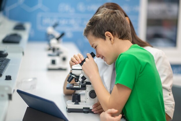 Junge, der durch das Mikroskop in der Nähe des sitzenden Lehrers schaut