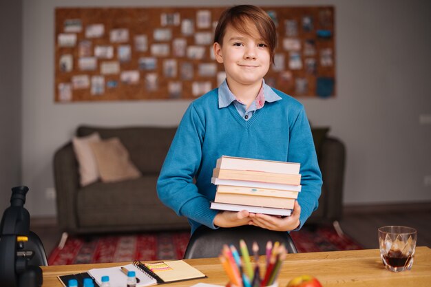 Junge der ersten Klasse, der zu Hause lernt, ein paar Bücher in der Hand hält und sich auf den Online-Unterricht vorbereitet