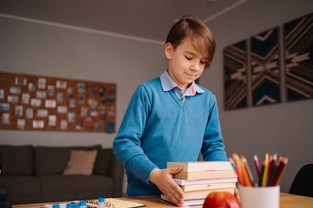 Junge der ersten Klasse, der zu Hause lernt, ein paar Bücher in der Hand hält und sich auf den Online-Unterricht vorbereitet