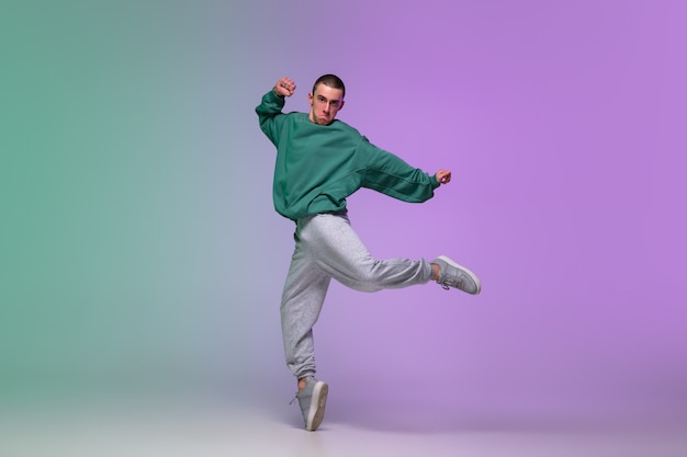 Junge, der Hip-Hop in stilvollen Kleidern auf Gradientenhintergrund an der Tanzhalle im Neonlicht tanzt.