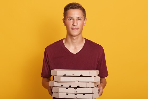 Junge, der kastanienbraunes lässiges T-Shirt trägt, das Pizzaschachteln liefert, lokalisiert auf Gelb posiert, Kamera betrachtet, sieht ernst aus, junge Frau, die als Lieferbote arbeitet und seine Arbeit tut. Personenkonzept