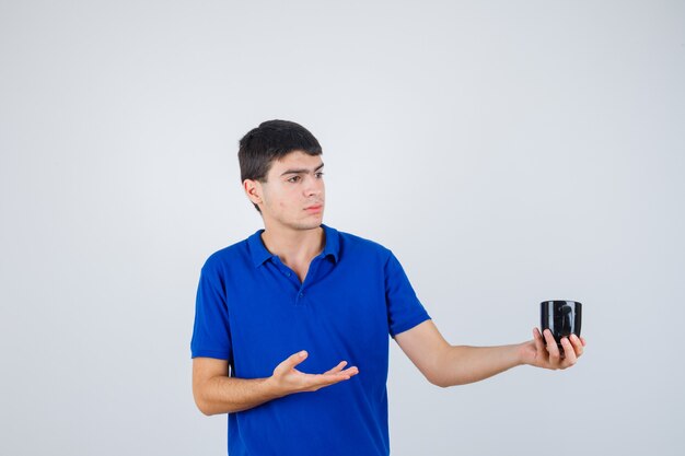 Junge, der Tasse hält, Hand dazu in blauem T-Shirt streckend und ernst aussehend