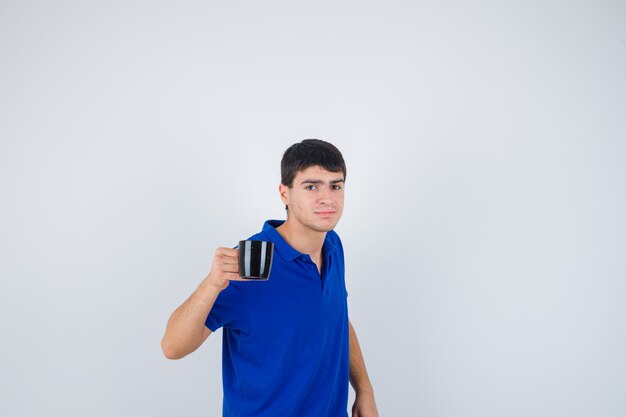 Junge, der Tasse hält, im blauen T-Shirt lächelnd und glücklich schaut, Vorderansicht.