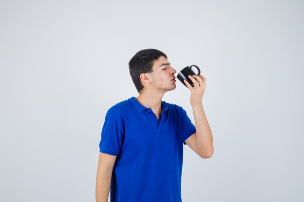 Junge, der Tasse hält, versucht, Flüssigkeit davon in blauem T-Shirt zu trinken und ernst aussehend