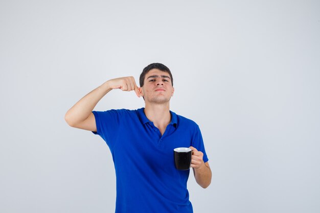 Junge, der Tasse nahe Kinn hält, geballte Faust im blauen T-Shirt und selbstbewusst aussehend, Vorderansicht.