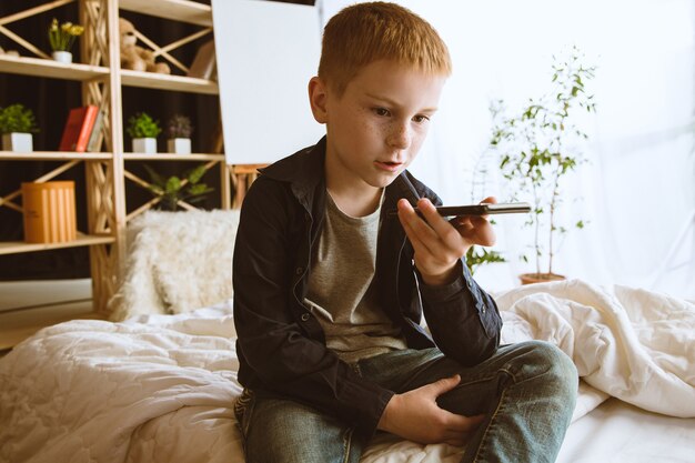 Junge, der zu Hause verschiedene Geräte verwendet. Kleines Modell mit Smartwatches, Smartphone oder Tablet und Kopfhörern. Selfie machen, chatten, spielen, Videos ansehen. Interaktion von Kindern und modernen Technologien.