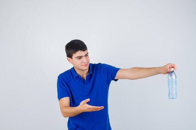 Junge, die Wasserflasche hält, Hand streckt, als sie es im blauen T-Shirt präsentiert und glücklich schaut. Vorderansicht.