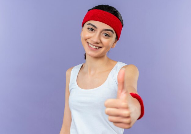 Junge Eignungsfrau in der Sportbekleidung mit dem Stirnband lächelnd, das fröhlich zeigt Daumen hoch, die über lila Wand stehen