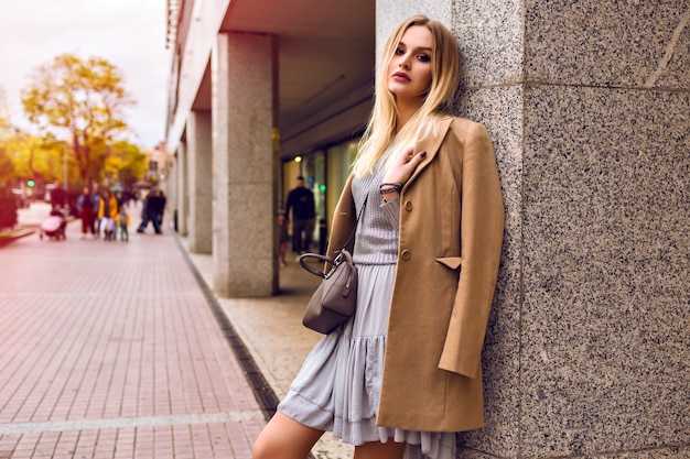 Junge elegante Frau, die auf der Straße nahe Einkaufszentrum, glamouröse trendige Outfits, beige Mantel, silberner Pullover und Kleid, Frühlingszeit, natürliche Schönheit aufwirft