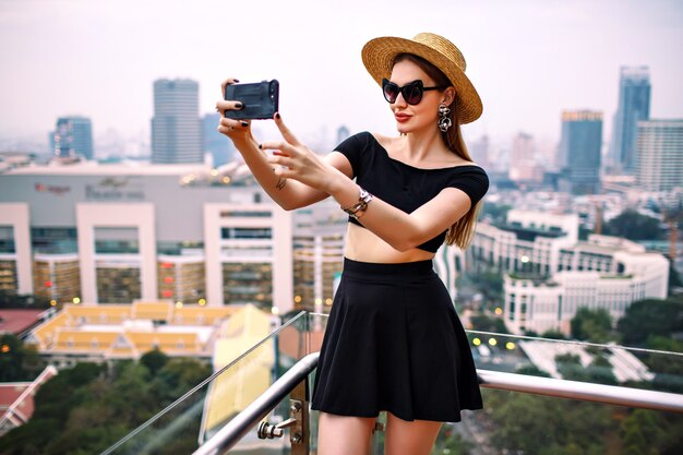 Junge elegante Frau, die modisches trendiges Sommeroutfit trägt, das touristisches Selfie an der Terrasse des Luxushotels macht