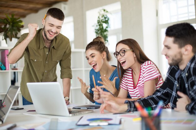 Junge emotionale Geschäftsleute, die freudig zusammen am Laptop arbeiten Gruppe lachender Männer und Frauen, die Zeit bei der Arbeit in einem modernen, gemütlichen Büro verbringen