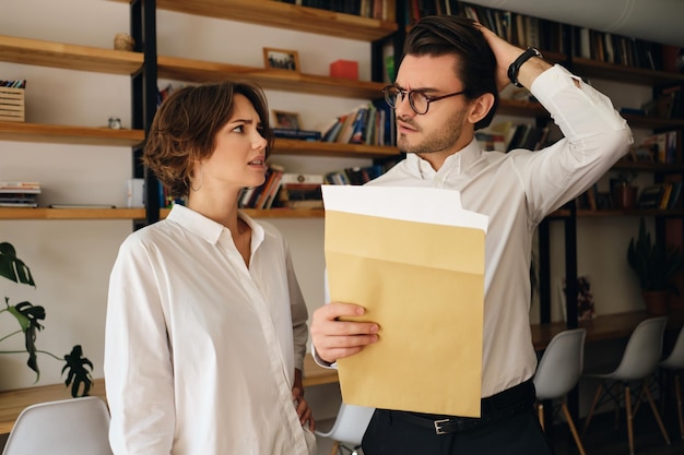 Junge enttäuschte Geschäftskollegen, die mit Briefumschlag stehen, während sie sich im modernen Büro traurig ansehen