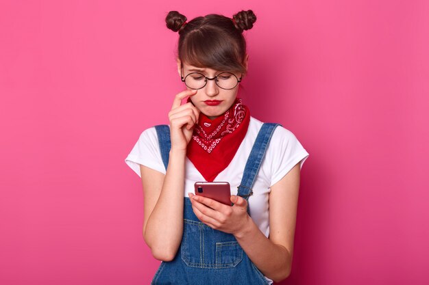 Junge entzückende Frau trägt Jeansoverall, lässiges weißes T-Shirt, rotes Kopftuch am Hals und abgerundete Brille, mit Trauben fühlt sich traurig an, während sie SMS auf ihrem Handy liest, isoliert über rosa