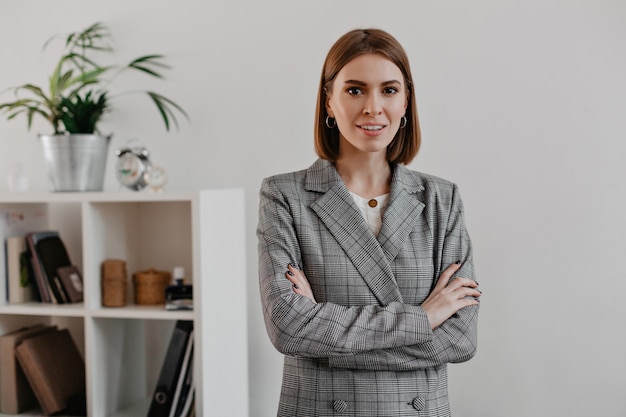 Kostenloses Foto junge erfolgreiche frau mit den braunen augen in der klassischen grauen jacke, die im hellen büro aufwirft.