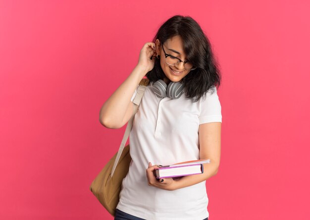 Junge erfreut hübsches kaukasisches Schulmädchen, das Brillentasche und Kopfhörer trägt, hält und betrachtet Buch und Notizbuch auf Rosa mit Kopienraum