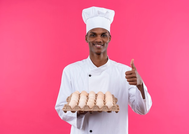 Junge erfreute afroamerikanische Köchin in der Kochuniform hält Charge von frischen Eiern und Daumen lokalisiert auf rosa Wand