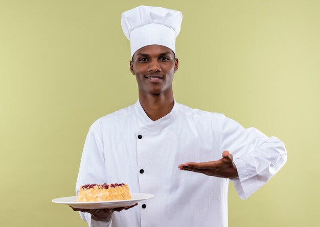 Junge erfreute afroamerikanische Köchin in der Kochuniform hält Kuchen auf Teller und zeigt mit Hand lokalisiert auf grüner Wand