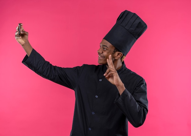Junge erfreute afroamerikanische Köchin in der Kochuniform schaut auf Telefon und gestikuliert Siegeshandzeichen lokalisiert auf rosa Wand