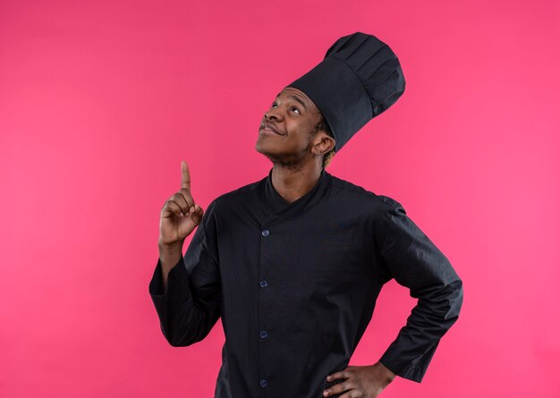 Junge erfreute afroamerikanische Köchin in der Kochuniform zeigt nach oben und legt Hand auf Taille isoliert auf rosa Wand