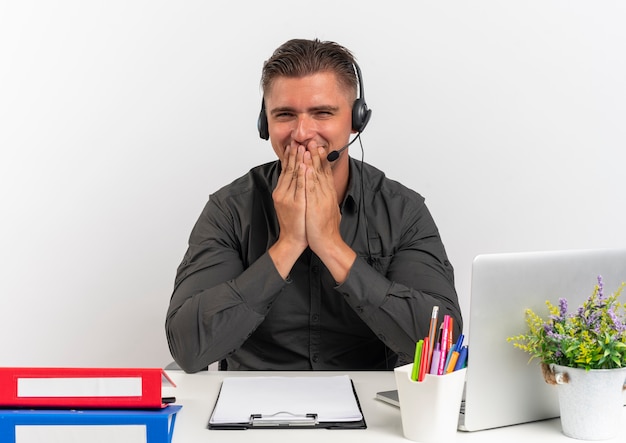 Junge erfreute blonde Büroangestellte Mann auf Kopfhörern sitzt am Schreibtisch mit Bürowerkzeugen unter Verwendung von Laptop setzt Hände auf Mund