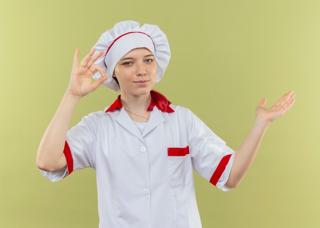 Junge erfreute blonde Köchin in Kochuniform gestikuliert ok Handzeichen und zeigt seitlich isoliert auf grüne Wand