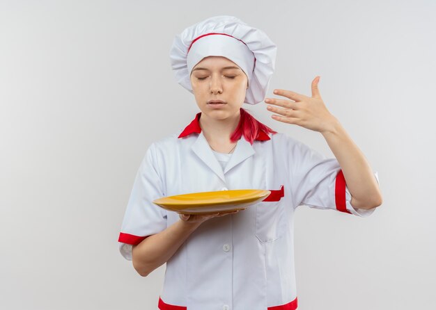 Junge erfreute blonde Köchin in Kochuniform hält Teller und gibt vor, isoliert auf weißer Wand zu riechen