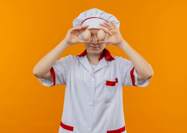 Junge erfreute blonde Köchin in Kochuniform schließt Augen mit Eiern, die auf orange Wand lokalisiert werden