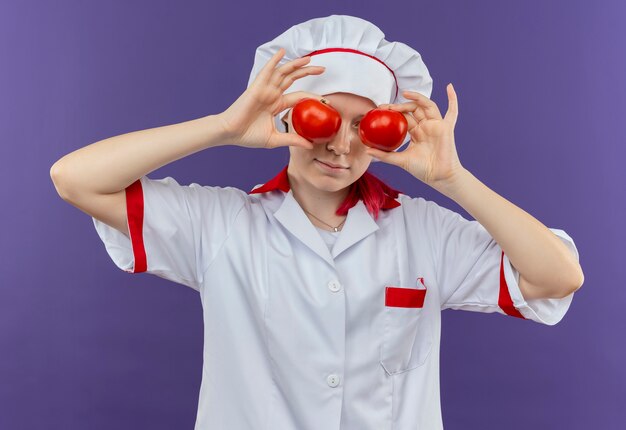 Junge erfreute blonde Köchin in Kochuniform schließt Augen mit Tomaten, die auf violetter Wand isoliert werden