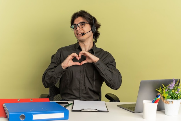 Junge erfreute büroangestellte mann auf kopfhörern in optischen gläsern sitzt am schreibtisch mit bürowerkzeugen unter verwendung von laptop-gesten-herzhandzeichen lokalisiert auf grünem hintergrund mit kopienraum