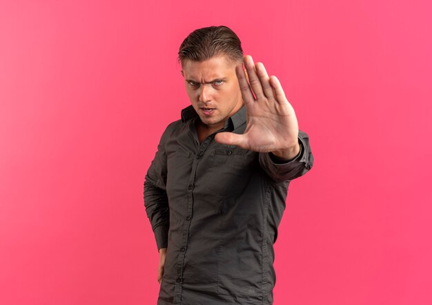 Junge ernsthafte blonde hübsche Manngesten stoppen Handzeichen lokalisiert auf rosa Hintergrund mit Kopienraum