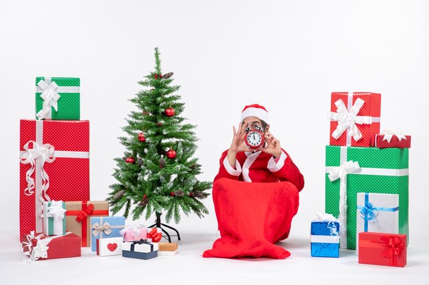 Junge Erwachsene feiern Weihnachtsfeiertage, die im Boden sitzen und Uhr nahe Geschenken und geschmücktem Weihnachtsbaum halten