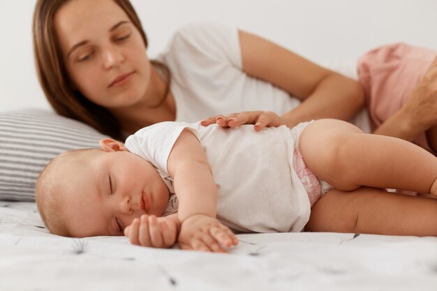 Junge erwachsene Frau mit dunklem Haar, die mit Baby im Bett liegt, Tochter anschaut, um sie schlafen zu sehen oder nicht, Frau mit weißem Casual-T-Shirt, glückliche Mutterschaft.