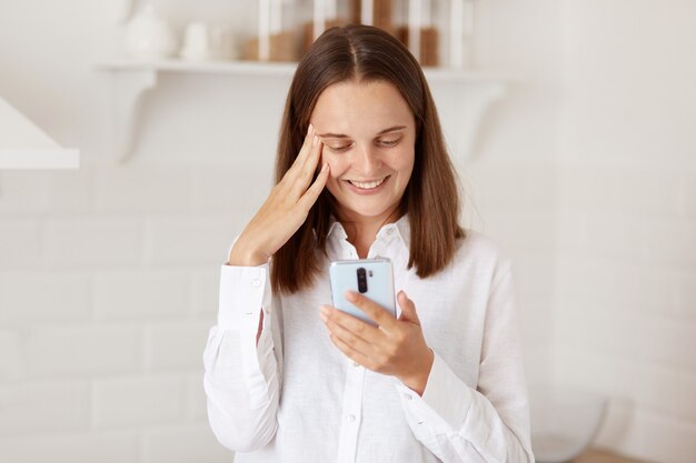 Junge erwachsene hübsche glückliche Frau, die Smartphone verwendet, mit Handy in der hellen Küche stehend, Finger an ihren Schläfen hält, lächelt, bewegliches Display betrachtend.