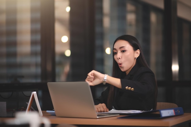 Junge erwachsene intelligente asiatische Geschäftsfrau im schwarzen Freizeitanzug mit Laptop, der Überstunden im städtischen Büro macht