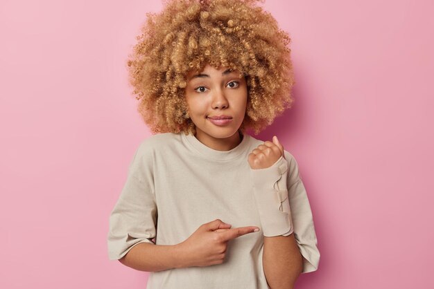 Junge Europäerin mit lockigem Haar zeigt gebrochenen Arm nach einem Unfall trägt einen elastischen Verband in einem lässigen T-Shirt, das über rosafarbenem Studiohintergrund isoliert ist Versicherungsunfall- und Gesundheitskonzept