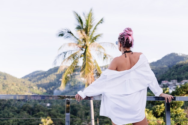 Junge europäische Frau im schwarzen Bikini des weißen langen Hemdes mit rosa Zöpfen auf Kopf auf tropischen Palmen