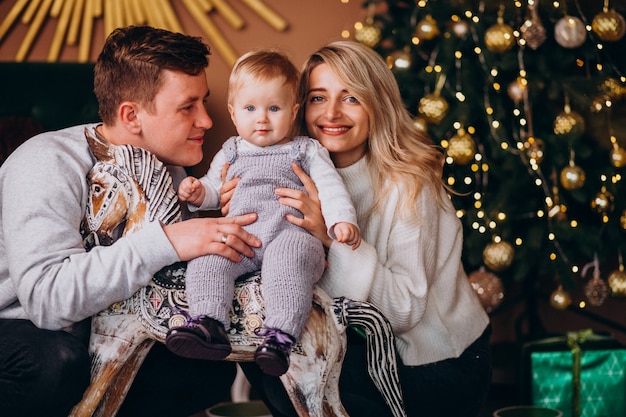 Junge Familie mit dem Baby, das durch Weihnachtsbaum sitzt