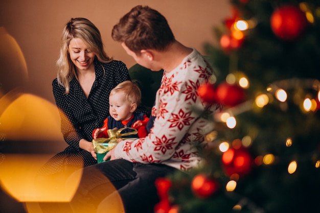 Junge familie mit dem baby, das durch weihnachtsbaum sitzt