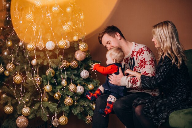 Junge Familie mit dem Baby, das durch Weihnachtsbaum sitzt