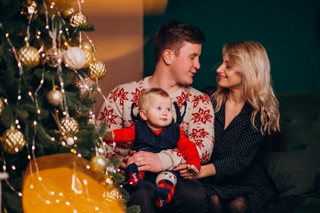 Junge Familie mit dem Baby, das durch Weihnachtsbaum sitzt