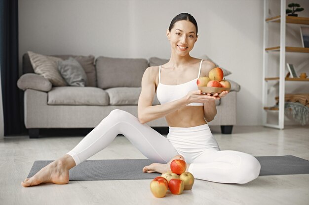 Junge Fitness-Frau sitzt zu Hause auf Yogamatte und hält frische Äpfel