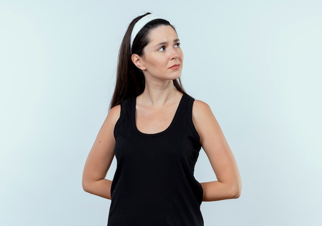 Junge Fitnessfrau im Stirnband, das beiseite mit nachdenklichem Ausdruck auf Gesicht steht, das über weißem Hintergrund steht