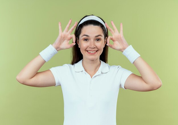 Junge Fitnessfrau im Stirnband, das Kamera betrachtet, die fröhlich zeigt ok Zeichen, das über hellem Hintergrund steht