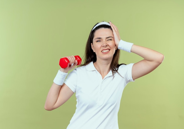 Junge Fitnessfrau im Stirnband, das mit der Hantel arbeitet, die verwirrt steht über heller Wand steht