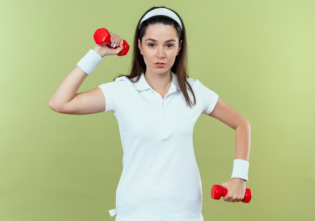 Junge Fitnessfrau im Stirnband, das mit Hanteln arbeitet, die Kamera mit ernstem Gesicht betrachten, das über hellem Hintergrund steht