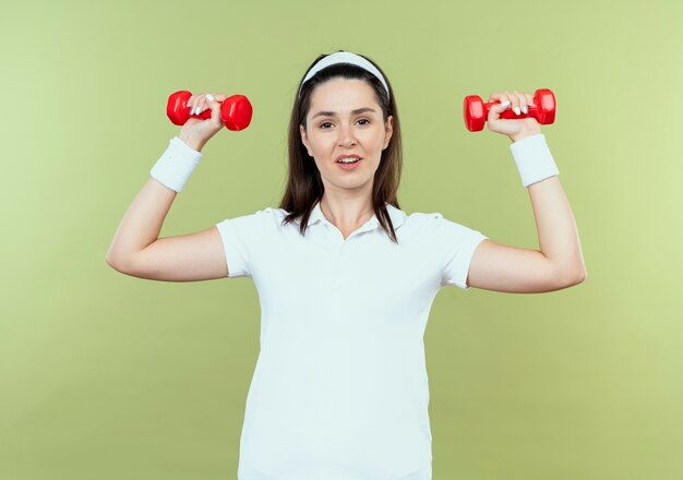 Junge Fitnessfrau im Stirnband, das mit Hanteln arbeitet, die sicher stehen über heller Wand stehen