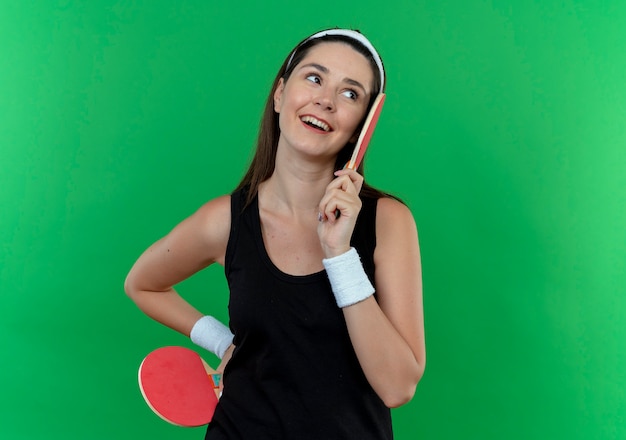 Junge Fitnessfrau im Stirnband, das Schläger für Tennistabelle hält, die beiseite lächelnd mit glücklichem Gesicht steht über blauem Hintergrund steht
