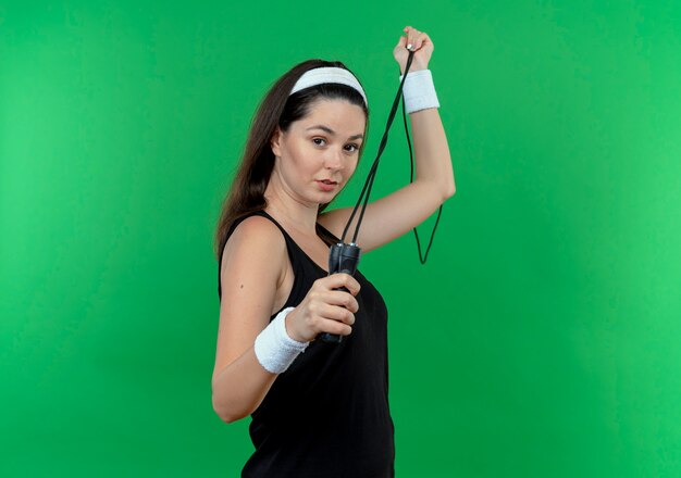 junge Fitnessfrau im Stirnband, das Springseil mit sicherem Ausdruck hält, der über grüner Wand steht