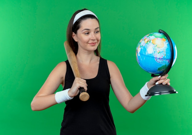 junge Fitnessfrau im Stirnband, die Baseballschläger und Globus hält, der es mit Lächeln auf Gesicht betrachtet, das über grüner Wand steht