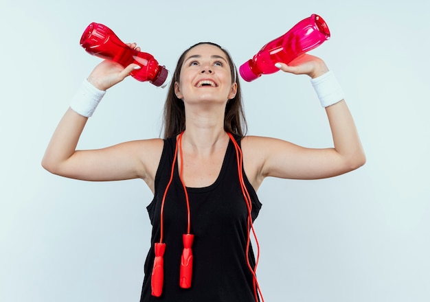 junge Fitnessfrau im Stirnband, die eine Flasche Wasser hält, die nach dem Training über der weißen Wand steht